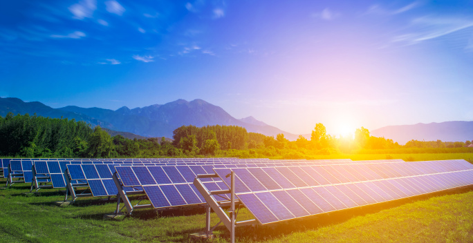 Auf diesem Bild erkennt man Photovoltaikanlagen auf einem Feld. Die Sonne scheint und im Hintergrund sieht man Berge.