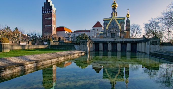 Historische Gebäude mit einem Park und Wasser im Vordergrund bei blauem Himmel