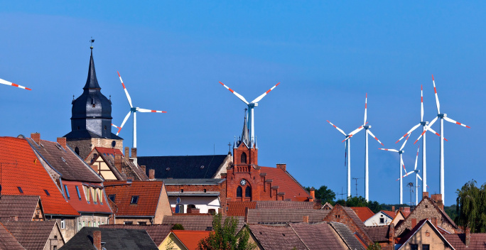 Das Bild zeigt eine Stadtlandschaft mit historischen Gebäuden und roten Ziegeldächern, über der sich mehrere moderne Windräder mit rot-weißen Flügelspitzen erheben.