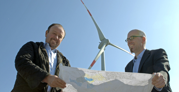 Zwei Männer stehen mit einer Karte vor einer Windkraftanlage.