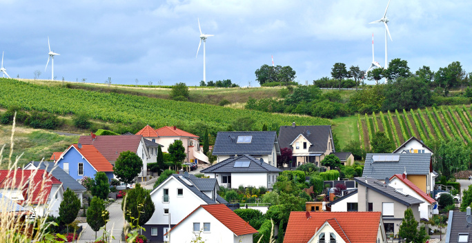 Im Vordergrund des Bildes stehen moderne Wohnhäuser mit Solarpanels. Dahinter erstrecken sich grüne Felder und mehrere Windräder.