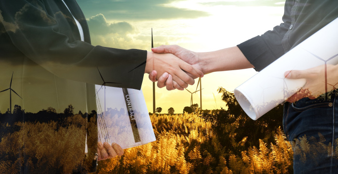 Auf dem Bild geben sich 2 Personen einen Handschlag. Im Hintergrund befinden sich Windräder in einer sonnigen Landschaft.