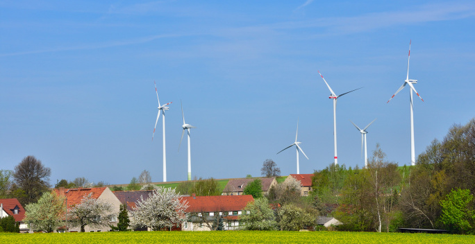 Hinter einer Wiese befindet sich ein kleines Dorf. Im Hintergrund drehen sich mehrere Windräder.