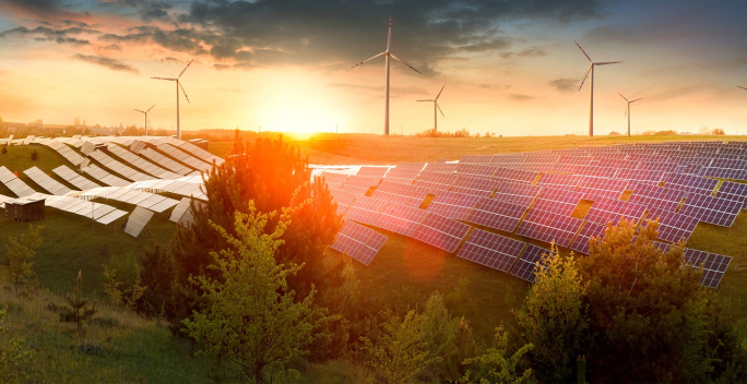 Im Vordergrund des Bild steht ein Solarpark auf einer hügeligen Landschaft. Dahinter befinden sich mehrere Windräder bei einem Sonnenuntergang.