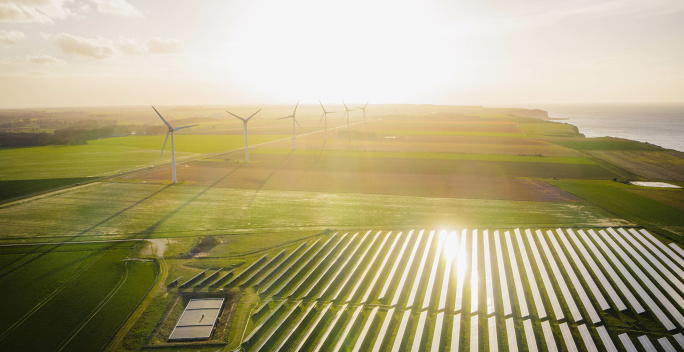 Das Bild zeigt eine grüne Landschaft mit Solarpaneelen im Vordergrund und Windkraftanlagen im Hintergrund, beleuchtet von warmem Sonnenlicht.