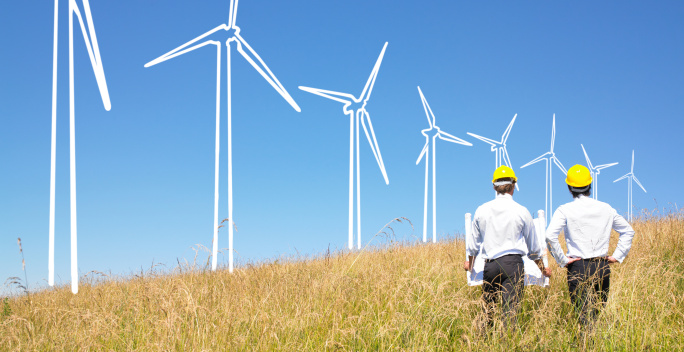 Windräder auf einem Feld, vor blauem Himmel, 2 Männer mit gelben Helmen stehen mit dem Rücken davor